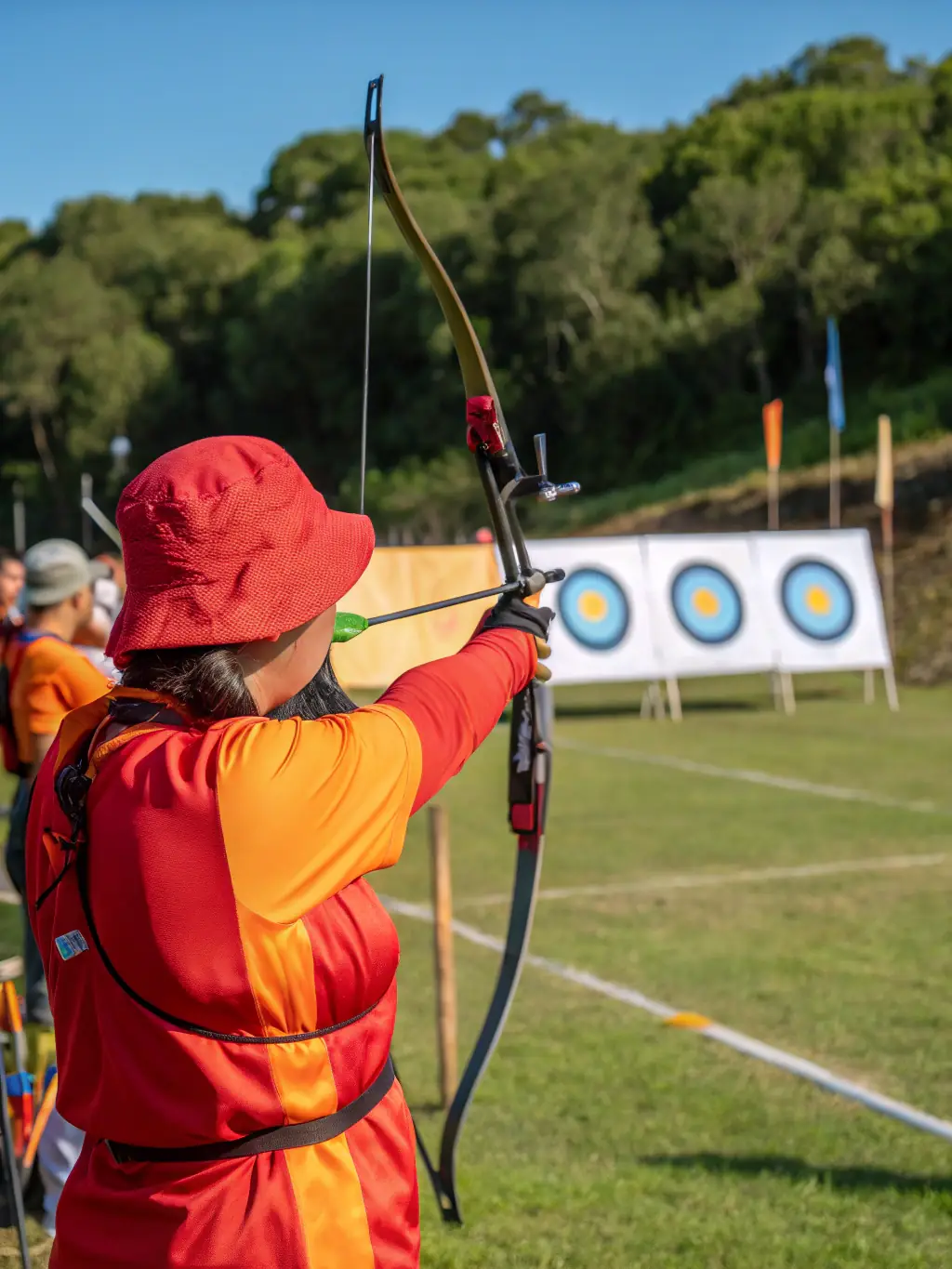 A focused shot of an archer drawing back their bow during a beginner's training session at Les Archers Embrunais, with other participants in the background.